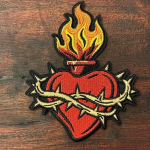 Puede incluir: Un parche bordado que muestra un corazón rojo rodeado por una corona de espinas dorada. Sobre el corazón, una llama estilizada en tonos amarillos y naranjas se eleva desde una pequeña base. Todo el diseño está delineado con hilo negro.