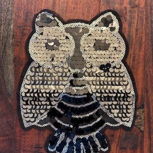 May include: A decorative owl applique made of silver and black sequins. The owl has large, round eyes and a detailed feather design. The applique is outlined with a black mesh and is suitable for embellishing clothing or accessories.