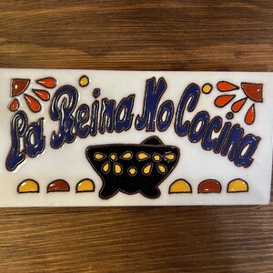 Peut inclure: Carreau de céramique blanc rectangulaire avec l'inscription "La Reina No Cocina" en bleu. Le carreau présente un bol noir avec des motifs de fleurs jaunes et des éléments décoratifs orange, jaune et rouge.