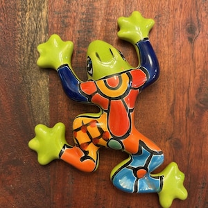 May include: A colorful ceramic frog figurine with a green head and feet, blue arms and legs, and a vibrant body design in orange, red, and yellow. The frog is posed in a playful, outstretched position.