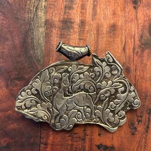 May include: A silver-toned metal belt buckle with an engraved deer design. The buckle features intricate floral and leaf patterns. A matching silver-toned metal piece is attached to the buckle. The buckle is displayed on a wooden surface.