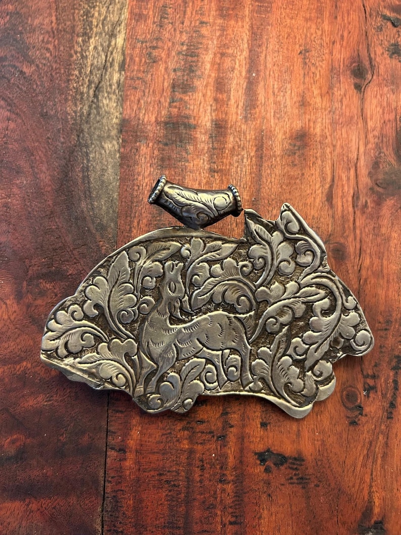 May include: A silver-toned metal belt buckle with an engraved deer design. The buckle features intricate floral and leaf patterns. A matching silver-toned metal piece is attached to the buckle. The buckle is displayed on a wooden surface.