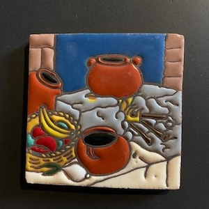 May include: A ceramic tile with a colorful painted design of three clay pots, fruit, and a wooden stove. The tile has a blue, white, and brown background.