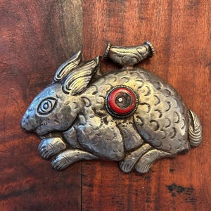 May include: A silver-toned metal rabbit figurine with detailed fur texture and a red and white circular accent. The rabbit has a small handle on its back. The figurine is lying on a wooden surface.