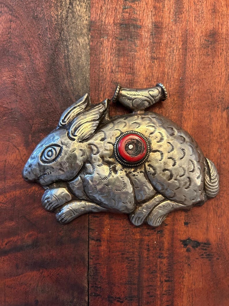 May include: A silver-toned metal rabbit figurine with detailed fur texture and a red and white circular accent. The rabbit has a small handle on its back. The figurine is lying on a wooden surface.