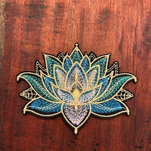 May include: Embroidered lotus flower patch featuring shades of blue, teal, and yellow. The patch has a black border and intricate stitching details. The lotus design is centered on a wooden surface.
