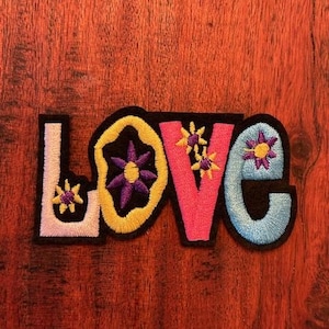 May include: Embroidered patch spelling "LOVE" in colorful, stylized letters. The "L" is white, the "O" is yellow with a purple flower, the "V" is pink with yellow flowers, and the "E" is light blue with a purple flower.