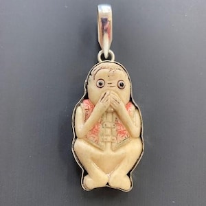 May include: A white carved pendant of a figure with its hands covering its eyes. The figure is wearing a pink and white patterned vest. The pendant is attached to a silver bail.