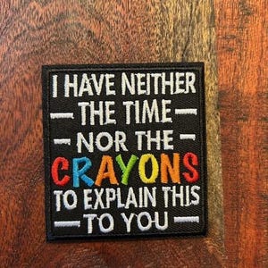 Puede incluir: Parche cuadrado negro con texto bordado en blanco: "I HAVE NEITHER THE TIME - NOR THE - CRAYONS TO EXPLAIN THIS - TO YOU". La palabra "CRAYONS" está en letras coloridas. El parche está sobre una superficie de madera.