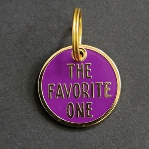 May include: A purple metal keychain with a gold ring and the text "THE FAVORITE ONE" in gold lettering.
