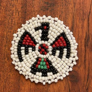 May include: A round beaded design with a black, white, red, and green color scheme. The design is centered on a white background.