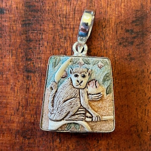 May include: Silver pendant with a detailed monkey design. The pendant features a monkey holding a banana, set against a colorful background. The pendant is rectangular with a decorative bail.
