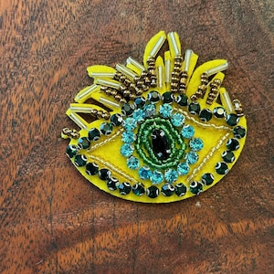 May include: A yellow felt eye patch with green and blue beaded details. The eye is surrounded by gold and silver beads.