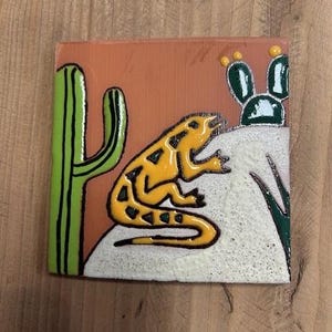 May include: A yellow and green ceramic tile featuring a cartoon lizard on a white background. The lizard is perched on a rock with a cactus and a green plant in the background.