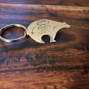 May include: Silver key chain with a bear design. The bear is stylized and has a detailed design on its back.