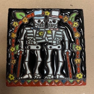 May include: A ceramic tile with a black background depicting two skeletons wearing top hats and holding canes. The skeletons are surrounded by a colourful floral garland. The tile has a red, orange, yellow, green and blue border.