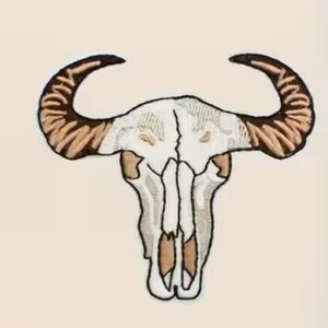 May include: Embroidered patch featuring a white bull skull with brown horns and tan accents. The skull has detailed stitching and is set against a neutral background. This patch is suitable for jackets, bags, or other fabric items.
