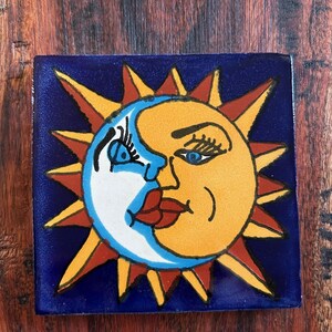 Puede incluir: Azulejo de cerámica cuadrado con un diseño celestial. La imagen representa un sol sonriente y una luna creciente, con ojos azules y labios rojos, sobre un fondo azul oscuro. El sol tiene rayos naranjas y rojos.