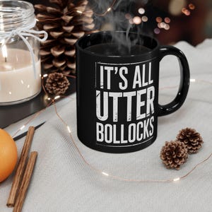 Puede incluir: Taza de cerámica negra con el texto "IT'S ALL UTTER BOLLOCKS" en blanco. La taza está llena de un líquido oscuro y sale vapor de la parte superior. La taza está sobre una superficie gris claro.