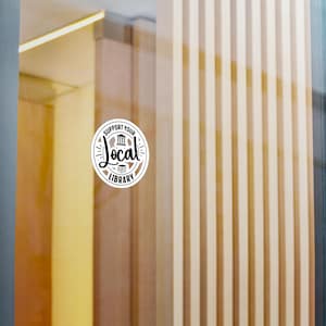Puede incluir: Una pegatina circular blanca aplicada sobre una superficie de cristal con el texto negro SUPPORT YOUR LOCAL LIBRARY, que incluye un pequeño icono de un edificio y una pila de libros dentro del diseño circular.