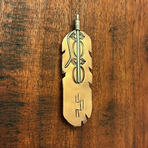 May include: A copper-colored pendant in the shape of a feather, with silver-colored accents. The pendant has a stylized design with a central rod and decorative elements. The bottom of the feather has etched symbols.