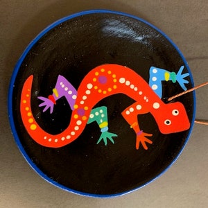May include: A hand-painted, decorative dish featuring a vibrant red lizard design with white polka dots and colorful accents. The lizard is set against a black background with a blue rim. The dish is likely used for decorative purposes.