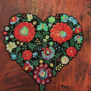 May include: A heart-shaped embroidered patch with a black background, featuring vibrant red, green, blue, and yellow floral designs. The patch is embellished with sequins and detailed stitching, creating a decorative and colorful accessory.