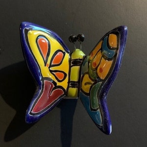 Puede incluir: Una decoración de pared de mariposa de cerámica colorida con acentos azules, amarillos, rojos, verdes y negros. La mariposa tiene un diseño detallado con patrones intrincados y un efecto tridimensional.