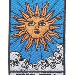 May include: A blue and white embroidered patch with a yellow sun with a face in the center. The sun is surrounded by rays and clouds. The text "THE SUN" is at the bottom of the patch.