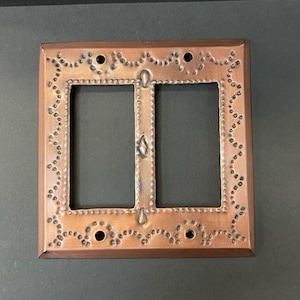 Könnte beinhalten: Eine kupferfarbene Doppel-Lichtschalterabdeckung. Die Abdeckung hat eine rechteckige Form mit einem dekorativen Rand aus kleinen, gleichmäßig verteilten Punkten und einem blütenartigen Design. Die Mitte hat zwei rechteckige Öffnungen.