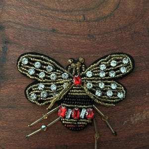 May include: A decorative bee-shaped patch featuring intricate beadwork in gold and black. The wings are adorned with clear rhinestones, while the body incorporates red rectangular crystals and gold seed beads. Long gold-toned beaded legs extend outward.