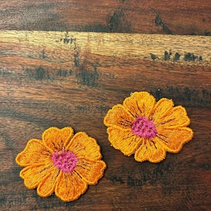 May include: Two embroidered flower patches featuring vibrant orange petals and a bright pink center. The floral designs are crafted with detailed stitching, showcasing a circular shape with distinct, rounded petal edges and a textured appearance.