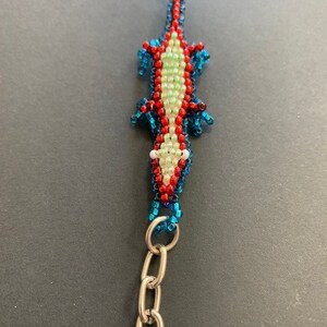 May include: A beaded keychain shaped like a blue and red lizard with a white belly. The lizard is attached to a silver chain.