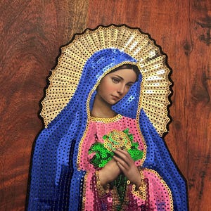 May include: A sequined patch featuring a portrait of the Virgin Mary. The image shows Mary with a blue veil, pink and gold dress, and a gold halo. She is holding a bouquet of green flowers. The patch is on a wooden surface.