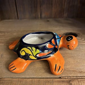 May include: A ceramic turtle-shaped planter with an orange body and head, and a dark blue shell decorated with colorful floral patterns. The planter has a white interior and is placed on a wooden surface.