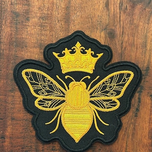 May include: Embroidered patch featuring a golden bee wearing a crown, set against a black background. The bee's wings and body are detailed with yellow thread, and the crown is intricately designed. The patch is likely for clothing or accessories.