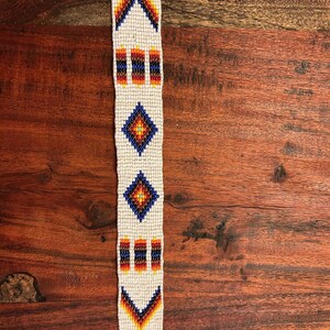 May include: A beaded strip with a cream background and geometric patterns in red, orange, yellow, and blue. The patterns include chevrons, rectangles, and diamond shapes. The strip is displayed on a wooden surface.