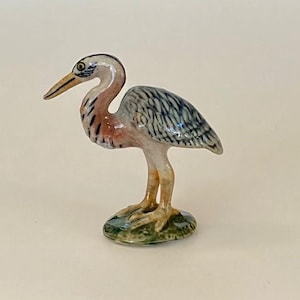 May include: A ceramic figurine of a heron, featuring a long, curved neck and beak. The bird has a detailed design with blue, gray, and brown feathers, standing on a green base. The heron's legs and beak are a light yellow color.