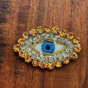 Puede incluir: Un amuleto de ojo turco blanco con un ojo azul y diamantes de imitación dorados y transparentes.