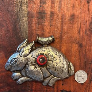 May include: A silver-colored metal rabbit figurine with intricate etched details. The rabbit has a red and white circular accent on its side. A small, decorative piece is attached to the top. A US quarter is next to the rabbit.