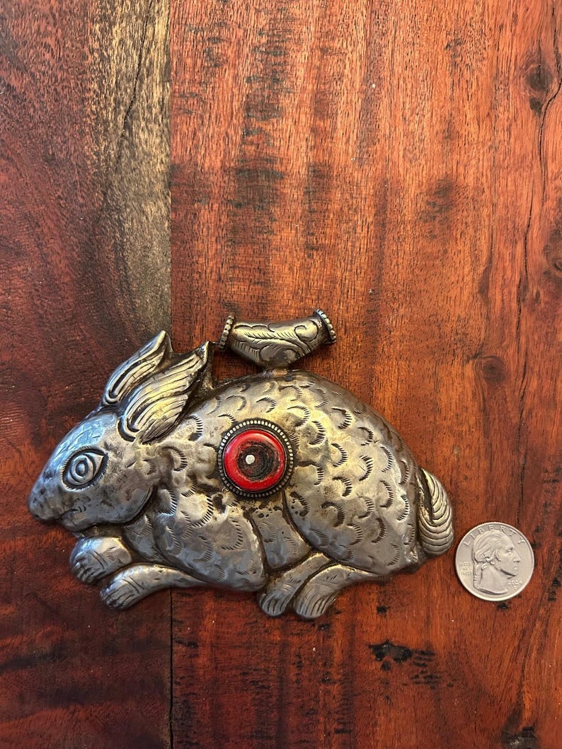 May include: A silver-colored metal rabbit figurine with intricate etched details. The rabbit has a red and white circular accent on its side. A small, decorative piece is attached to the top. A US quarter is next to the rabbit.