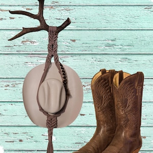 May include: A rustic scene featuring a cowboy hat hanging from a macrame holder attached to a decorative antler. A pair of brown leather cowboy boots with intricate stitching are placed beside the hat. The background is a weathered, turquoise-colored wooden wall.