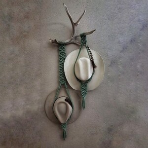 May include: Two straw cowboy hats hanging from an antler rack. The hats are both beige and have green macrame cord hanging from them.