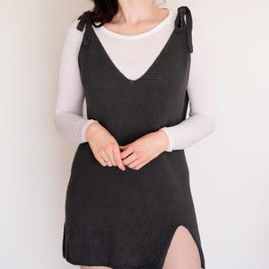 May include: A dark gray knitted dress with a V-neck and tie straps. The dress has a side slit and is worn over a white long-sleeved shirt. A white headband is also visible.