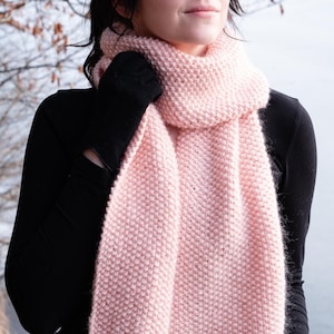 Könnte beinhalten: Ein weicher rosa gestrickter Schal mit einem strukturierten Muster. Der Schal wird von einer Person getragen, die ein schwarzes Hemd trägt.