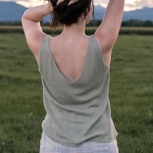 Könnte beinhalten: Ein hellgrünes Tanktop mit tiefem V-Ausschnitt. Die Person trägt ein hellgrünes Tanktop und eine karierte Hose. Die Person steht auf einem Feld mit Bergen im Hintergrund. Die Person hält sich die Haare.