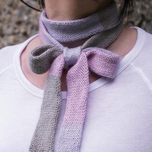 Könnte beinhalten: Ein gestrickter Schal in Grau-, Lavendel- und Rosatönen, zu einer Schleife gebunden. Der Schal ist um den Hals gewickelt und fällt nach vorne. Das weiche, strukturierte Material ist in der Nahaufnahme sichtbar. Der Schal ist ein Modeaccessoire.