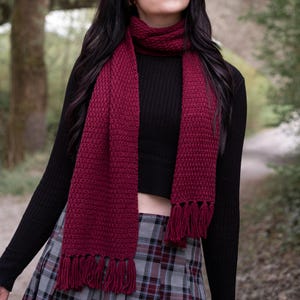 Puede incluir: Una bufanda de punto color burdeos con borlas de flecos. La bufanda se lleva alrededor del cuello de una persona que lleva una camisa negra de cuello alto y una falda de cuadros.
