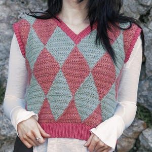 May include: A pink and green crochet argyle vest. The sleeveless top has a V-neck and a ribbed hem. The argyle pattern is made up of large diamond shapes. The vest is worn over a long-sleeved white shirt.