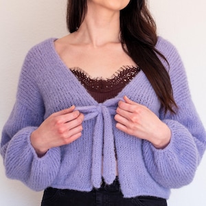 Puede incluir: Un cárdigan de punto de color lila claro con un frente de nudo y mangas largas. El cárdigan está hecho de un hilo suave y esponjoso y tiene un ajuste holgado.